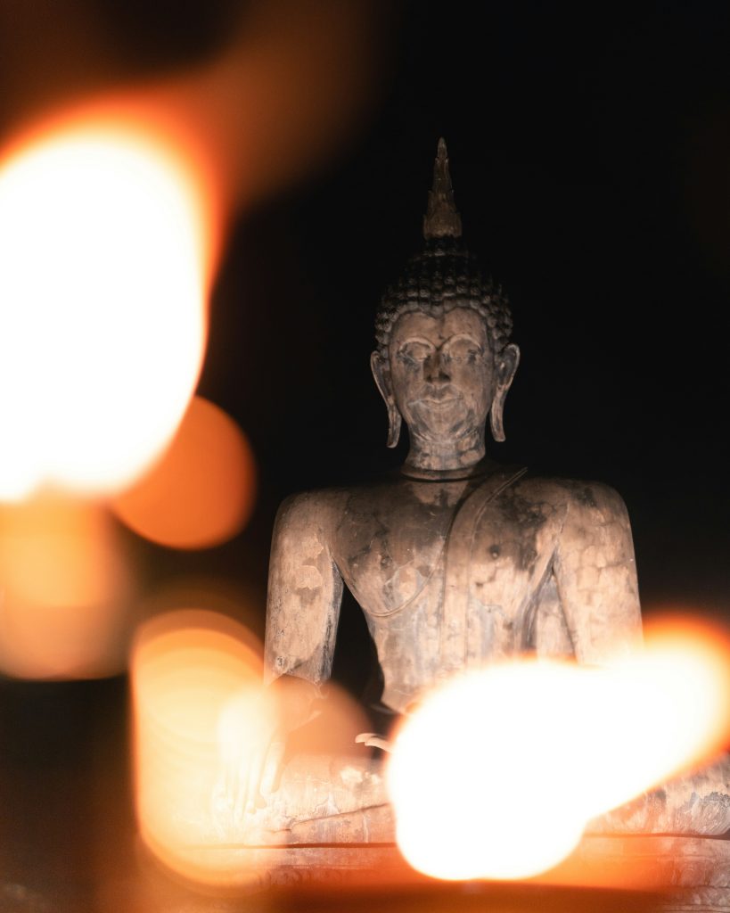 Seated Buddha statue in Thailand seen through soft candlelight bokeh in a dark temple setting.
