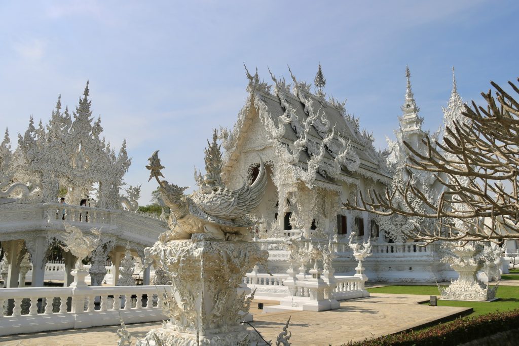 Wat Rong Khun (White Temple) in Chiang Rai, Thailand, with intricate white carvings and ornate sculptural details under a clear sky.
