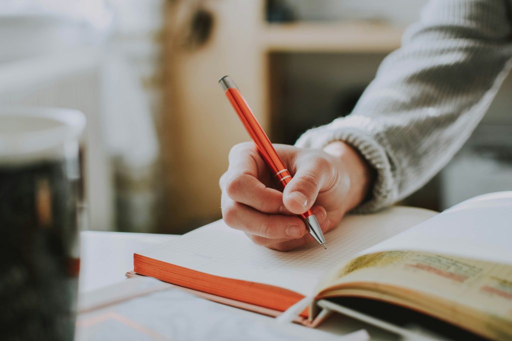 Person writing in a notebook with a red pen at a desk.