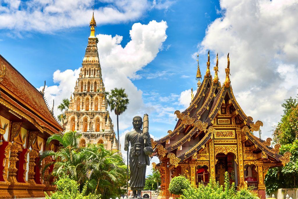 Ornate Thai temple complex with a tall chedi, golden Lanna-style roofs, a standing Buddha statue, and lush greenery under a bright blue sky.