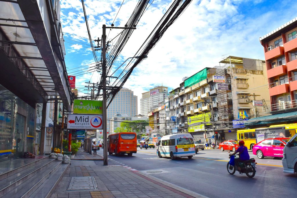 Busy street scene in Bangkok, Thailand, with traffic, colorful taxis, local buses, and overhead power lines on a sunny day.
