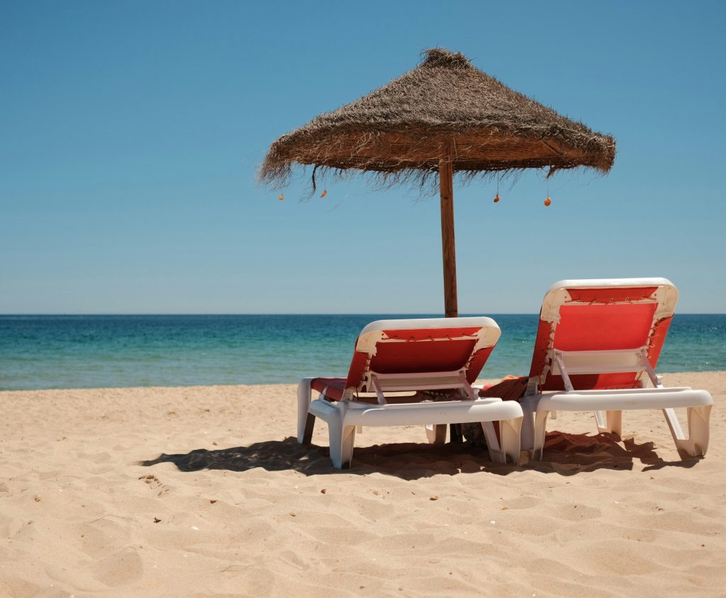Two sun loungers under a straw beach umbrella on a sandy shore by the sea.