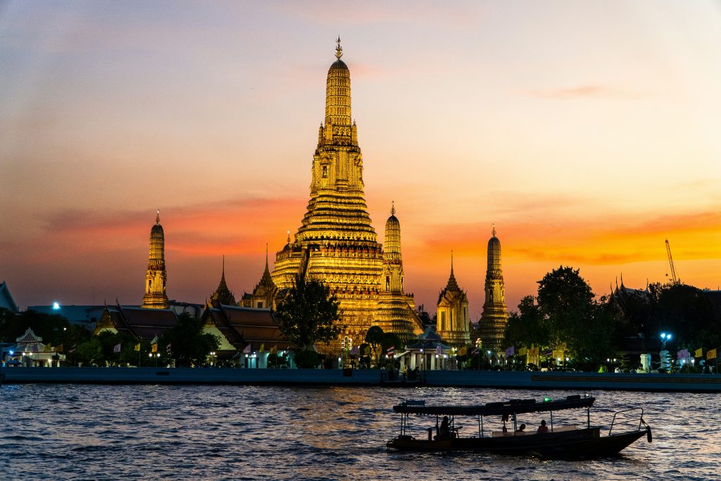 Wat Arun in Bangkok, Thailand, glowing at sunset across the Chao Phraya River with a boat in the foreground.